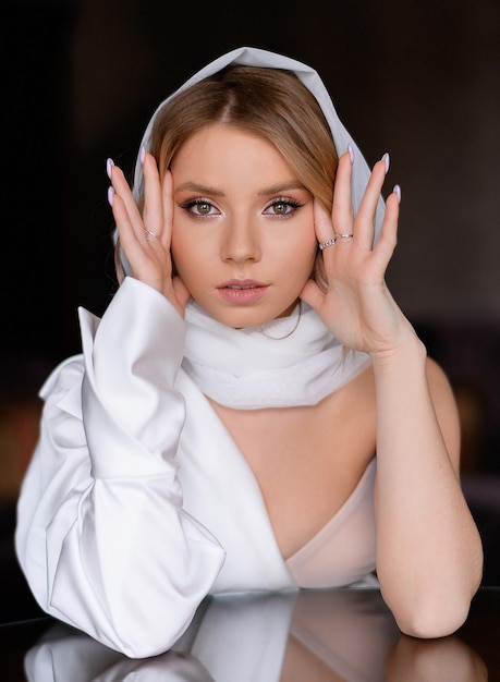 Retrato de una hermosa chica caucásica rubia con ojos marrones y bufanda blanca de seda en la mano Atractiva modelo femenina cogida de la mano cerca de la cara Apariencia de belleza Ropa elegante Maquillaje profesional desnudo