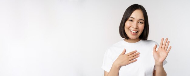 Retrato de una hermosa chica coreana levantando la mano para presentarse y poner la mano en el corazón saludando de pie sobre fondo blanco