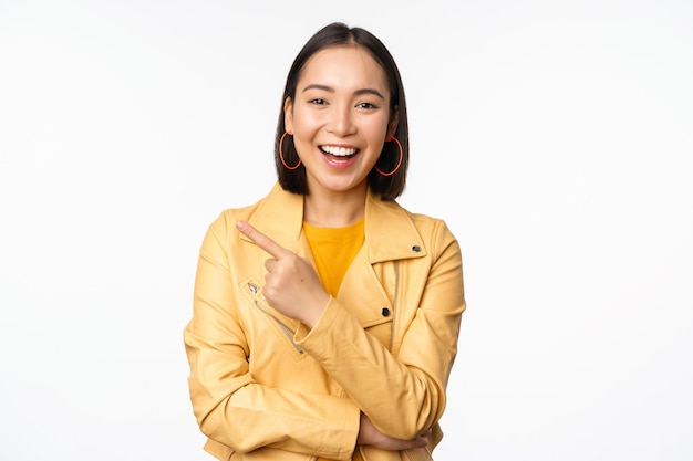 Retrato de una hermosa chica coreana riéndose sonriendo y señalando con el dedo a la izquierda en el logo que muestra una pancarta de pie con ropa informal sobre fondo blanco