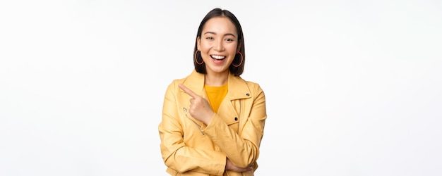 Retrato de una hermosa chica coreana riéndose sonriendo y señalando con el dedo a la izquierda en el logo que muestra una pancarta de pie con ropa informal sobre fondo blanco