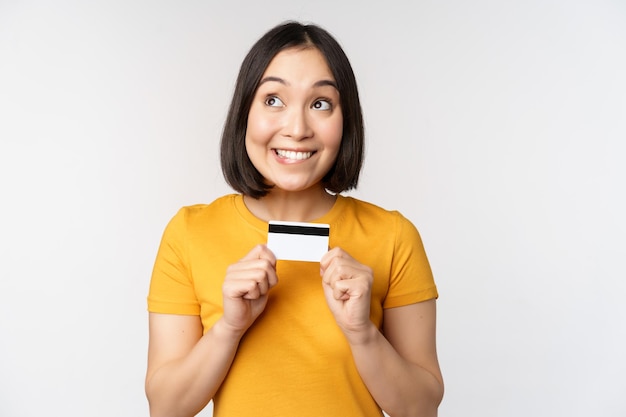 Retrato de una hermosa chica coreana sosteniendo una tarjeta de crédito recomendando el servicio bancario de pie en una camiseta amarilla sobre fondo blanco