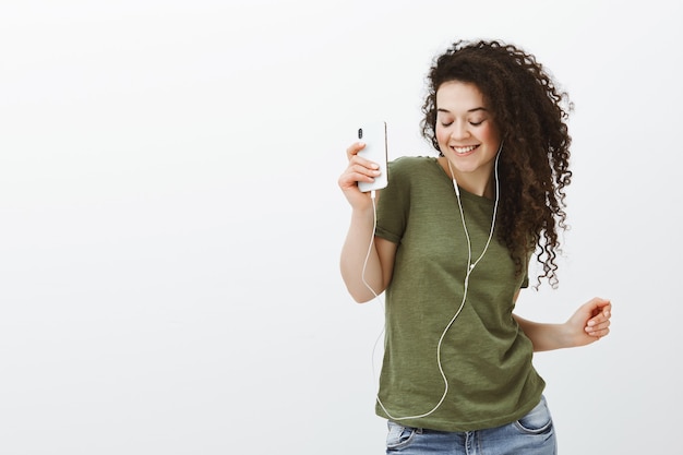 Retrato de una hermosa chica elegante sin preocupaciones con el pelo rizado, bailando con los ojos cerrados y una amplia sonrisa mientras sostiene el teléfono inteligente y escucha música en los auriculares