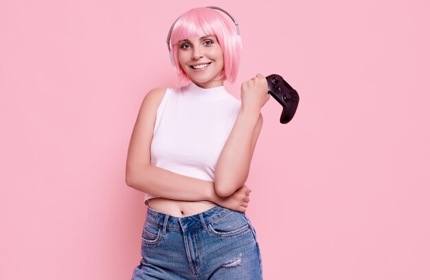 Retrato de hermosa chica gamer feliz con cabello rosado jugando videojuegos con joystick en colorido en estudio