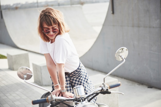 Foto gratuita retrato de una hermosa chica hipster sentada en un scooter retro negro, sonriendo posando y disfrutando del cálido sol primaveral.