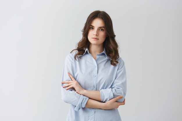 Retrato de hermosa chica impresionante con cabello oscuro cruzando las manos con expresión relajada y seria posando para el álbum de graduación universitaria.