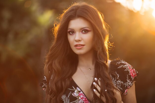 Retrato de una hermosa chica morena con cabello grueso y ondulado contra una puesta de sol de la ciudad Una chica con pantalones vaqueros cortos y una blusa negra colorida con maquillaje elegante y joyas