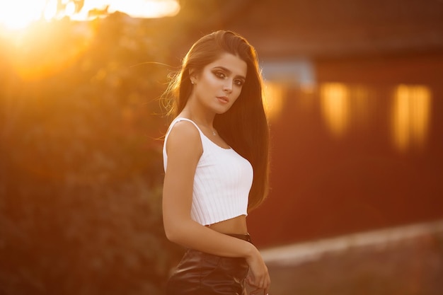 Retrato de una hermosa chica morena con cabello grueso ondulado sobre un fondo borroso de una puesta de sol de la ciudad Mujeres en una falda de cuero y una camiseta blanca con un elegante maquillaje Primer plano