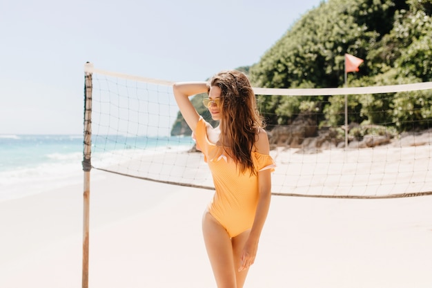 Retrato de hermosa chica morena de pie junto al juego de voleibol. Foto al aire libre de hermosa mujer bronceada en traje de baño naranja esperando el juego en la playa.
