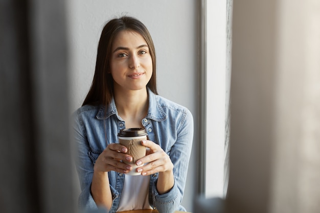 Foto gratuita retrato de hermosa chica relajada con cabello oscuro en camiseta blanca debajo de la camisa vaquera sonriendo mientras bebe una taza de café.