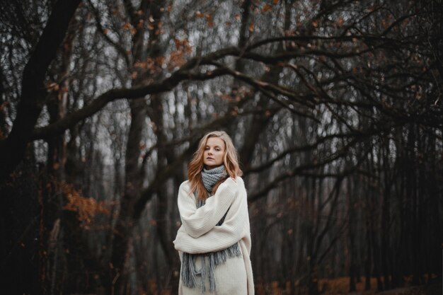 Retrato de hermosa chica rubia caucásica vestida con jersey blanco, curling solo en el bosque de otoño