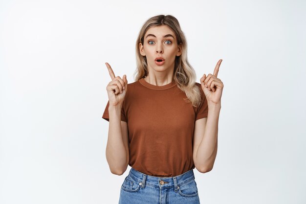 Retrato de hermosa chica rubia mirando sorprendido, apuntando hacia los lados de pie sobre blanco