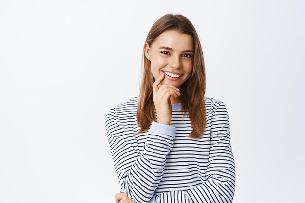 Retrato de una hermosa chica rubia que tiene una idea interesante escuchándote mirando a la cámara y sonriendo complacido de pie pensativo contra el fondo blanco