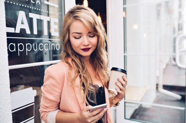 Retrato hermosa chica rubia en la terraza con una taza de café. Viste chaqueta coral, labios vinosos, sonriendo al teléfono en la mano.