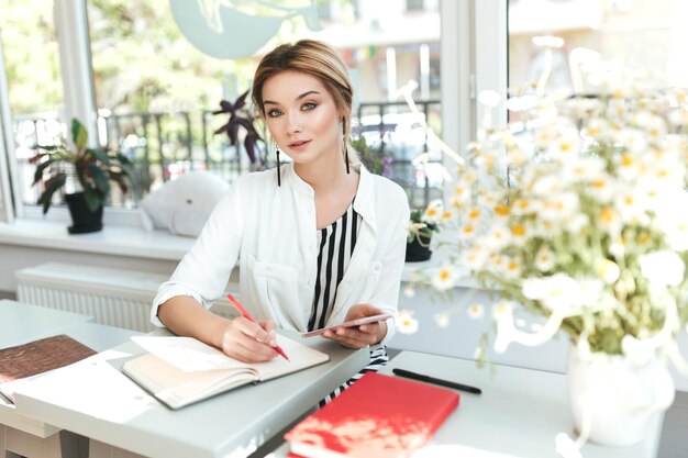 Retrato de una hermosa chica sentada en un restaurante con un cuaderno y mirando a puerta cerrada. Chica guapa con cabello rubio sosteniendo el teléfono móvil en la mano y escribiendo notas en su cuaderno en el café