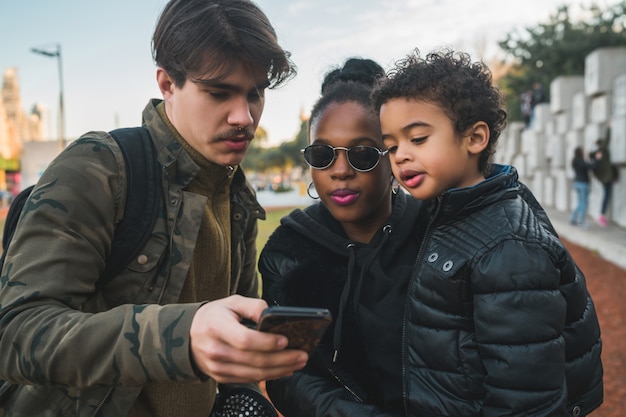 Retrato de hermosa familia étnica de raza mixta divirtiéndose, relajándose y usando el teléfono móvil en el parque al aire libre.