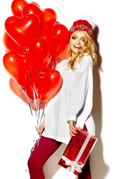 Retrato de hermosa feliz dulce mujer rubia sonriente niña sosteniendo en sus manos una gran caja de regalo de Navidad y globos de corazón en ropa casual de invierno hipster rojo, en suéter blanco cálido