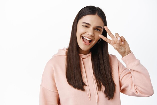 Retrato de una hermosa y feliz modelo femenina que se mantiene positiva guiñando un ojo y mostrando la paz contra el fondo blanco con capucha