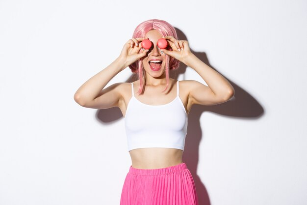 Retrato de hermosa fiestera con peluca rosa, sosteniendo macarrones sobre los ojos y sonriendo asombrado, de pie sobre fondo blanco.