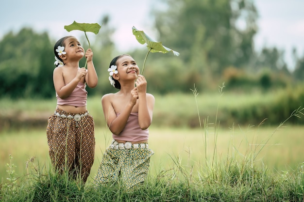 Retrato de hermosa hermana y hermana joven en traje tradicional tailandés y poner flor blanca en su oreja, mirando a la hoja de loto en la mano y sonriendo feliz en el campo de arroz, espacio de copia