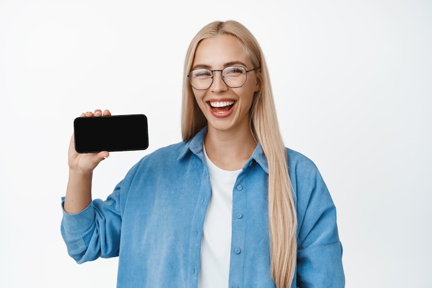 Retrato de una hermosa joven con anteojos sonriendo mostrando el teléfono inteligente horizontalmente la aplicación de teléfono móvil de pie sobre fondo blanco Copiar espacio