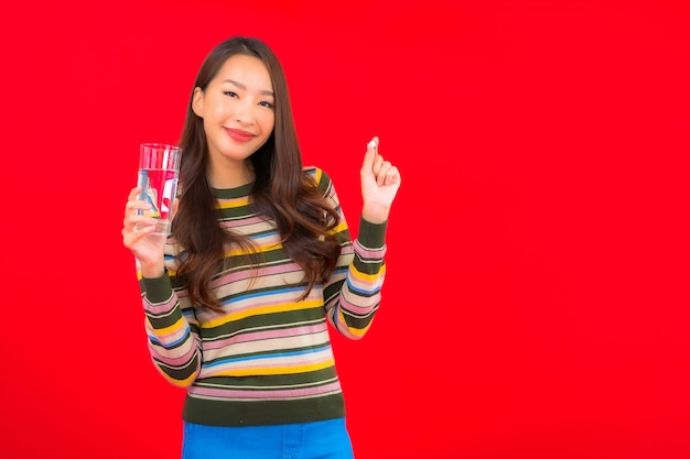 Retrato hermosa joven asiática con agua potable y píldora en la pared roja