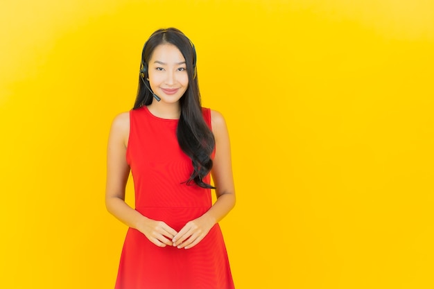 Retrato hermosa joven asiática con auriculares para comunicación y servicio de centro de llamadas de apoyo en la pared amarilla