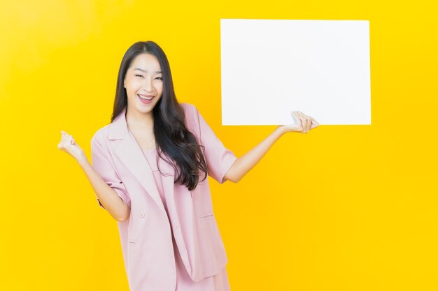 Retrato hermosa joven asiática con cartelera blanca vacía en la pared amarilla