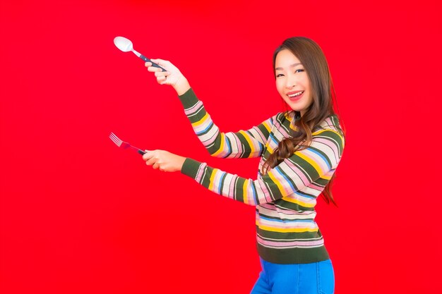 Retrato hermosa joven asiática lista para comer con cuchara y tenedor en la pared roja