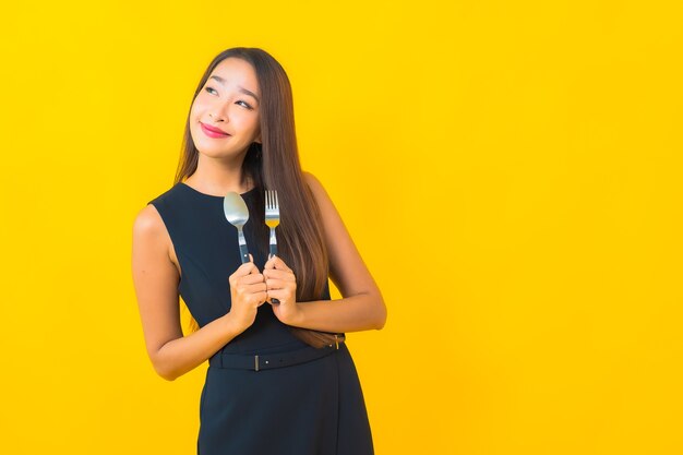 Retrato hermosa joven asiática lista para comer con tenedor y cuchara sobre fondo amarillo