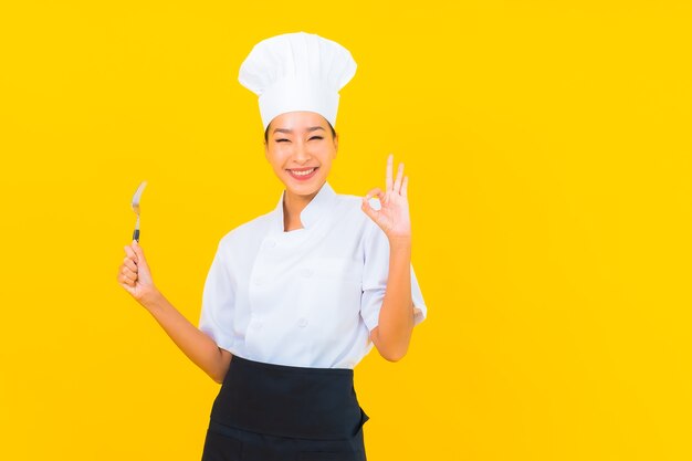 Retrato hermosa joven asiática llevar uniforme de cocinero o cocinero con cuchara y tenedor sobre fondo amarillo aislado