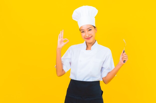 Retrato hermosa joven asiática llevar uniforme de cocinero o cocinero con cuchara y tenedor sobre fondo amarillo aislado