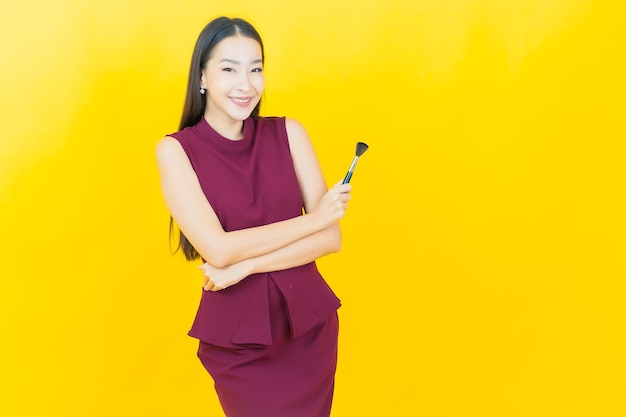 Retrato hermosa joven asiática con maquillaje cepillo cosmético en pared amarilla