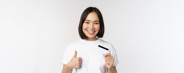 Retrato de una hermosa joven asiática moderna que muestra la tarjeta de crédito y el pulgar hacia arriba recomendando el pago sin contacto de pie sobre fondo blanco