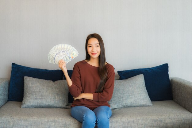 Retrato hermosa joven asiática mostrar dinero en efectivo y dinero en el sofá en el interior de la sala de estar