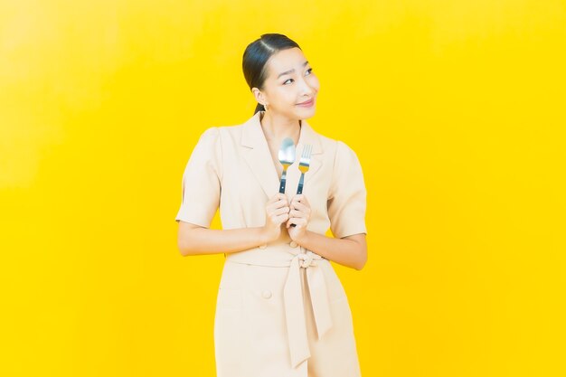Retrato hermosa joven asiática sonríe con cuchara y tenedor en la pared de color