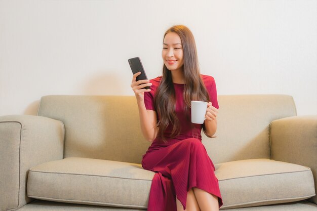 Retrato hermosa joven asiática con teléfono móvil inteligente en el sofá en el interior de la sala de estar