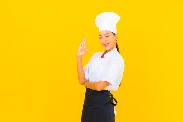 Retrato hermosa joven asiática en uniforme de cocinero o cocinero con sombrero sobre fondo amarillo aislado