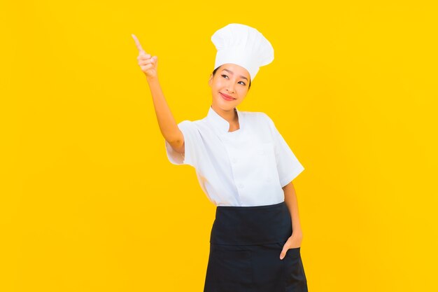 Retrato hermosa joven asiática en uniforme de cocinero o cocinero con sombrero sobre fondo amarillo aislado