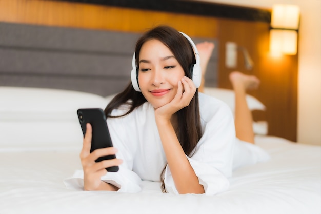 Retrato hermosa joven asiática usa teléfono móvil inteligente con auriculares para escuchar música en la cama en el interior del dormitorio