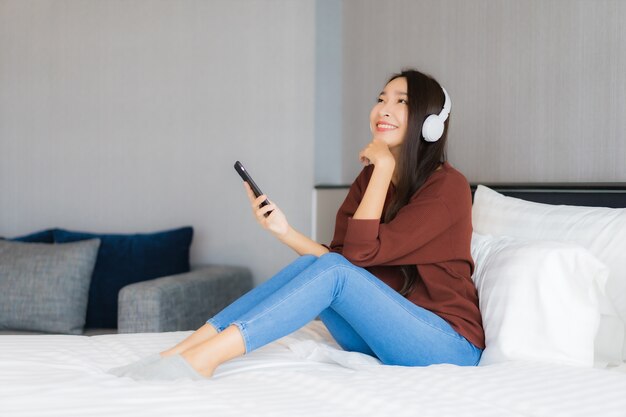 Retrato hermosa joven asiática usa teléfono móvil inteligente con auriculares para escuchar música en la cama en el interior del dormitorio