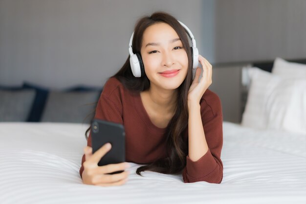 Retrato hermosa joven asiática usa teléfono móvil inteligente con auriculares para escuchar música en la cama en el interior del dormitorio