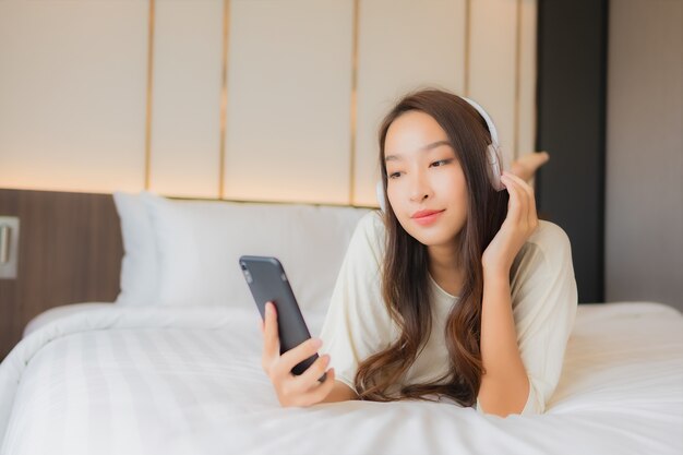 Retrato hermosa joven asiática usa teléfono móvil inteligente con auriculares para escuchar música en el dormitorio