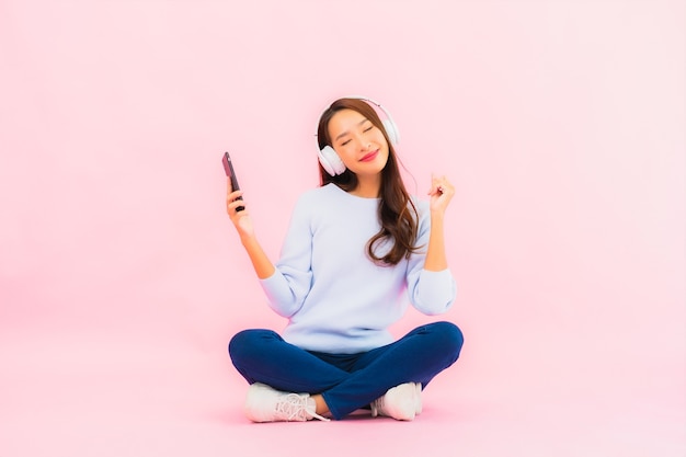 Retrato hermosa joven asiática usa teléfono móvil inteligente con auriculares para escuchar música en la pared rosa