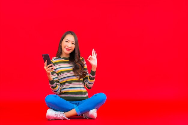 Retrato hermosa joven asiática usa teléfono móvil inteligente en pared roja aislada