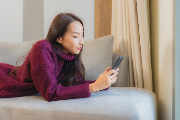 Retrato hermosa joven asiática usa teléfono móvil inteligente en el sofá en el interior de la sala de estar