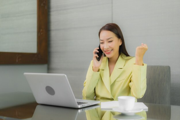 Retrato hermosa joven asiática usar computadora portátil con teléfono móvil inteligente en la mesa de trabajo en la habitación interior