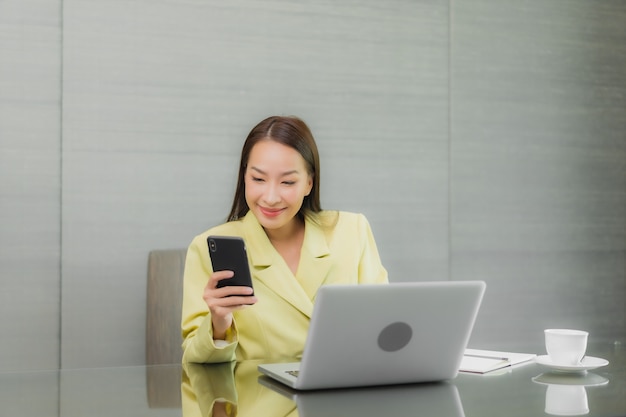 Retrato hermosa joven asiática usar computadora portátil con teléfono móvil inteligente en la mesa de trabajo en la habitación interior