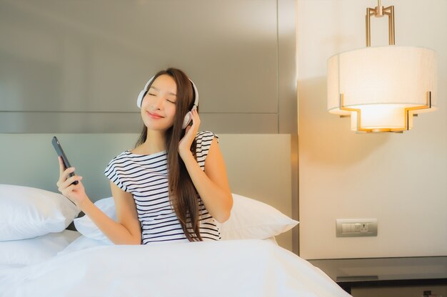 Retrato hermosa joven asiática utiliza teléfono móvil inteligente con auriculares para escuchar música en el interior del dormitorio