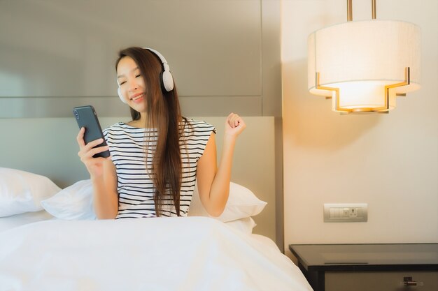 Retrato hermosa joven asiática utiliza teléfono móvil inteligente con auriculares para escuchar música en el interior del dormitorio