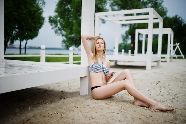 Retrato de una hermosa joven en bikini sentada junto al gasebo en la arena y posando